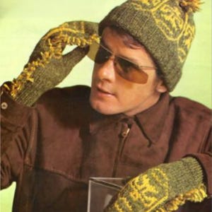 Puede incluir: Un hombre con una chaqueta marrón, un gorro de punto verde y amarillo con un pompón y guantes de punto a juego. Lleva gafas de sol.