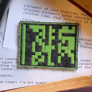 May include: A close-up of a black and green embroidered patch with a pixelated design. The patch is on a white piece of paper with text that reads "Statement of Jane Prentiss, regarding the original statement given February..."