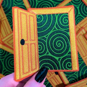 May include: A yellow and orange embroidered patch with a door opening to reveal a green background with a swirling pattern. The patch is held in a hand with black nail polish.