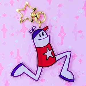 Puede incluir: Un llavero caprichoso con un personaje de dibujos animados. El personaje tiene una camisa roja con una estrella blanca, un gorro morado y piernas blancas. Está unido a un dije dorado en forma de estrella y un cierre dorado. El fondo es de color rosa claro con brillos en forma de estrella.