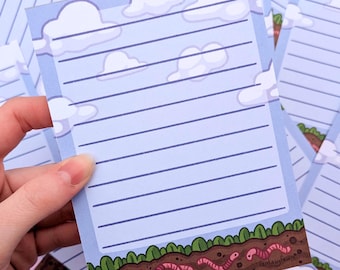 May include: A light blue notepad with horizontal lines, white clouds, and a green and brown border with cartoon worms. The notepad is held in a hand. The notepad has the text "Fantasy Fawn".