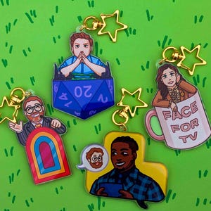 Peut inclure: Collection de porte-clés en acrylique colorés avec des fermoirs en forme d'étoile dorée. Les porte-clés présentent des illustrations de personnes, un dé D20, une arche arc-en-ciel et une tasse à café avec le texte "FACE FOR TV".