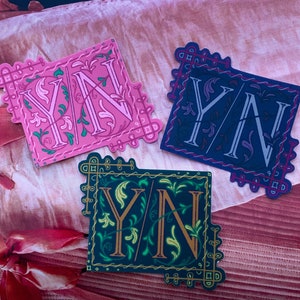 Può includere: Tre adesivi decorativi con le lettere "YN" in un font stilizzato. Gli adesivi sono rosa, blu e verdi con accenti dorati.