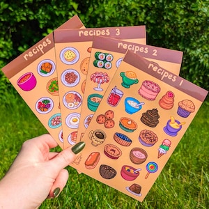 May include: Four recipe cards with illustrated food items, including sushi, cake, and soup. The cards are labeled "recipes 1", "recipes 2", and "recipes 3". The cards are held in front of a green background.