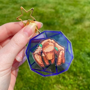 May include: A clear acrylic keychain featuring a brown crab illustration with green accents, set against a geometric blue and clear background. The keychain has a gold star-shaped clasp.