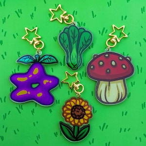 May include: A selection of novelty keyrings with colourful designs. The keyrings depict a purple star-shaped fruit, a green leafy vegetable, a red mushroom, and a yellow sunflower. Each charm is attached to a gold-coloured star-shaped clasp.