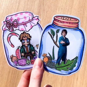 Puede incluir: Dos tarros de vidrio con tapas, decorados con diseños coloridos. Un tarro presenta un personaje de dibujos animados con caramelos y un bastón de caramelo. El otro tarro presenta a una persona con un mono azul y una planta.