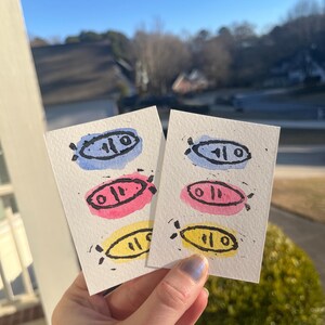 May include: Two small, rectangular watercolour paintings featuring fish designs in blue, pink, and yellow. Each fish has a black outline and details. The paintings are held up against a sunny outdoor background.