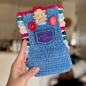 May include: A crochet pouch featuring a blue overall design with two red buttons and a patch that reads Good Guys. The pouch has a striped red, white, green, and blue background and includes a wooden tag with the text Wanna Play?