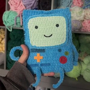 May include: A blue plush toy shaped like a rectangular video game console with a light green screen, a black smiling face, an orange cross-shaped button, a red circular button, a small green circular button, and thin blue arms and legs.