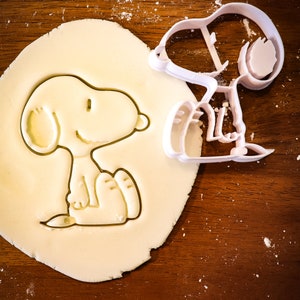 Könnte beinhalten: Ein weißer Snoopy-Ausstecher aus Kunststoff neben einem Snoopy-Ausstich in Teig. Der Teig liegt auf einer Holzoberfläche, die mit Mehl bestäubt ist. Der Ausstecher hat die Form von Snoopy, der sitzt, mit sichtbaren Armen und Beinen.