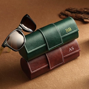 Puede incluir: Gafas de sol sobre dos estuches de cuero para gafas. El estuche superior es verde con costuras blancas e iniciales doradas "BB". El estuche inferior es burdeos con costuras blancas e iniciales doradas "AS". Las gafas tienen montura negra y patillas marrón claro. Los estuches están apilados.