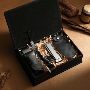 May include: A black gift box with a hinged lid, containing a black leather nail clipper set, a black leather strap, and a black leather case. The box is lined with straw-like material. A small jar and wood pieces are in the background.