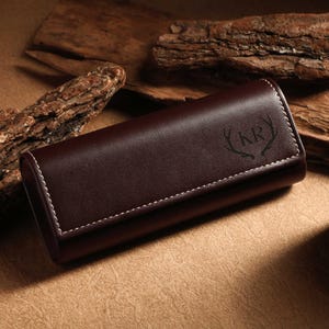 Puede incluir: Un estuche de cuero marrón oscuro con costuras blancas y un diseño de cornamenta en relieve con las iniciales "KR". El estuche rectangular está cerrado. El fondo incluye trozos de madera y una vela.