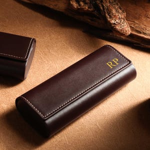 May include: A brown leather case with white stitching and gold initials "RP" on the front. Another similar case is in the background. The cases are on a brown surface.