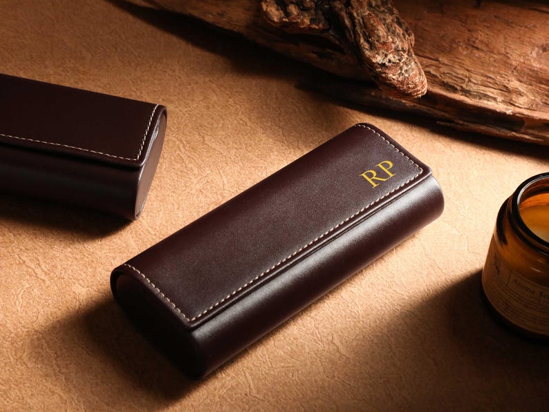 May include: A brown leather case with white stitching and gold initials "RP" on the front. Another similar case is in the background. The cases are on a brown surface.