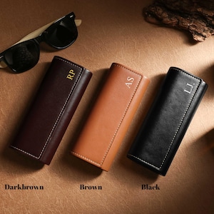 May include: Three leather eyeglass cases in dark brown, brown, and black, each with white stitching and personalized initials. Sunglasses rest above. The cases are labeled "Darkbrown", "Brown", and "Black".