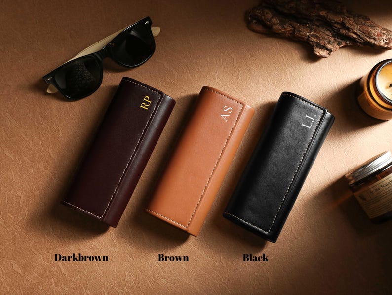 May include: Three leather eyeglass cases in dark brown, brown, and black, each with white stitching and personalized initials. Sunglasses rest above. The cases are labeled "Darkbrown", "Brown", and "Black".