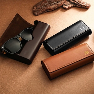 May include: Sunglasses with black frames and lenses rest in a dark brown leather case. Two additional leather cases, one black and one tan, are also present. The black case has the initials "L.J." and the tan case has "JR."