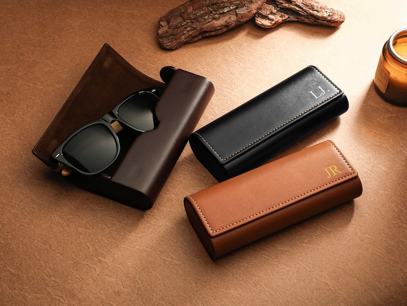 May include: Sunglasses with black frames and lenses rest in a dark brown leather case. Two additional leather cases, one black and one tan, are also present. The black case has the initials "L.J." and the tan case has "JR."