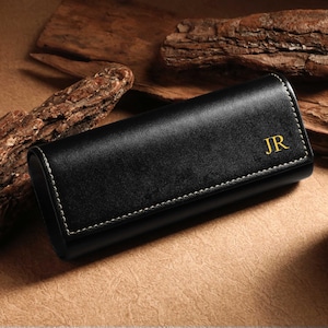 May include: Black leather glasses case with white stitching and gold initials "JR". The case is rectangular and closed, with a curved top. The background includes wood pieces and a brown surface.