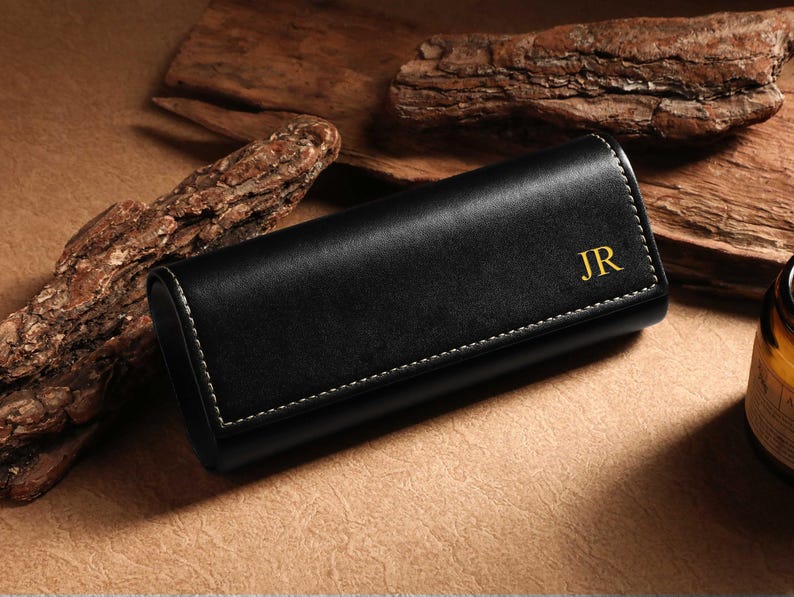 May include: Black leather glasses case with white stitching and gold initials "JR". The case is rectangular and closed, with a curved top. The background includes wood pieces and a brown surface.