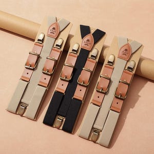 Puede incluir: Tres pares de tirantes exhibidos sobre cartón. Dos pares son de color beige con detalles de cuero marrón y herrajes color latón. El tercer par es negro con detalles de cuero marrón y herrajes color latón. Cada par tiene una etiqueta de cuero personalizada.