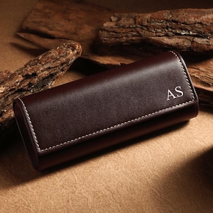 May include: A dark brown leather watch case with white stitching and the initials "AS" in the corner. The case is rectangular and closed, resting on a textured surface with wooden accents and a candle.