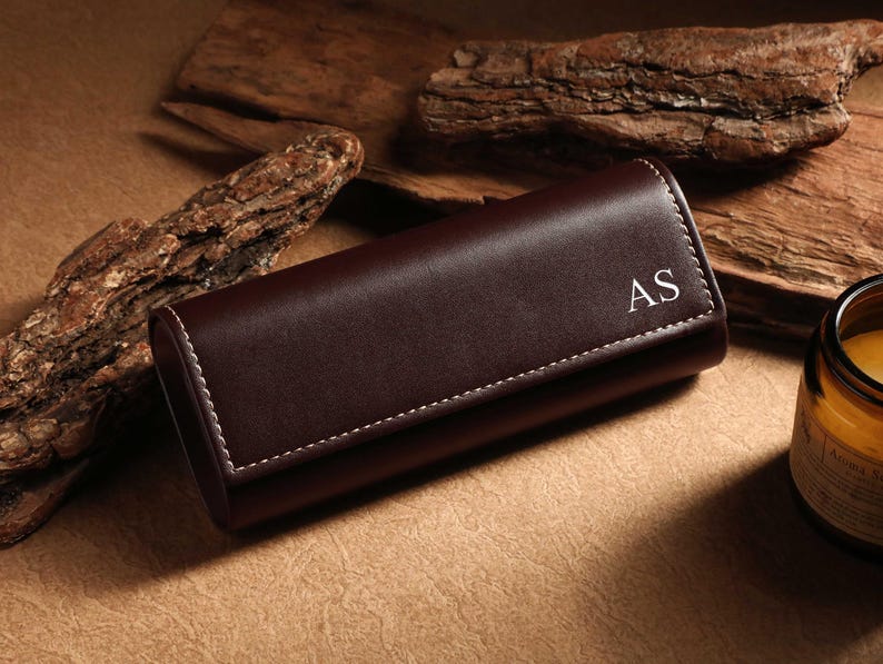 May include: A dark brown leather watch case with white stitching and the initials "AS" in the corner. The case is rectangular and closed, resting on a textured surface with wooden accents and a candle.