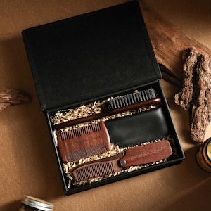 Puede incluir: Una caja de regalo negra que contiene herramientas de aseo. La caja incluye un peine de madera, un cepillo, una cartera de cuero negro y un peine pequeño. Los artículos están colocados sobre papel dorado. Una vela y trozos de madera están al lado.
