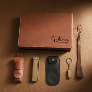 Puede incluir: Un juego de polipiel marrón con una caja rectangular con el nombre "Wilhelm", una armónica, un llavero, una funda de cuero y una correa de muñeca. Los artículos están dispuestos sobre una superficie marrón.
