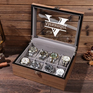 Peut inclure: Un coffret en bois pour montres avec un couvercle en verre, contenant dix montres. L'intérieur est en velours gris et le nom "Alexander" est gravé sur le verre. Les montres ont différents bracelets et cadrans métalliques.