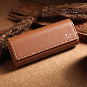 May include: A brown leather glasses case with white stitching and the initials "L.J." in white. The case is rectangular with rounded edges and a flap closure. The background includes wood pieces and a candle.