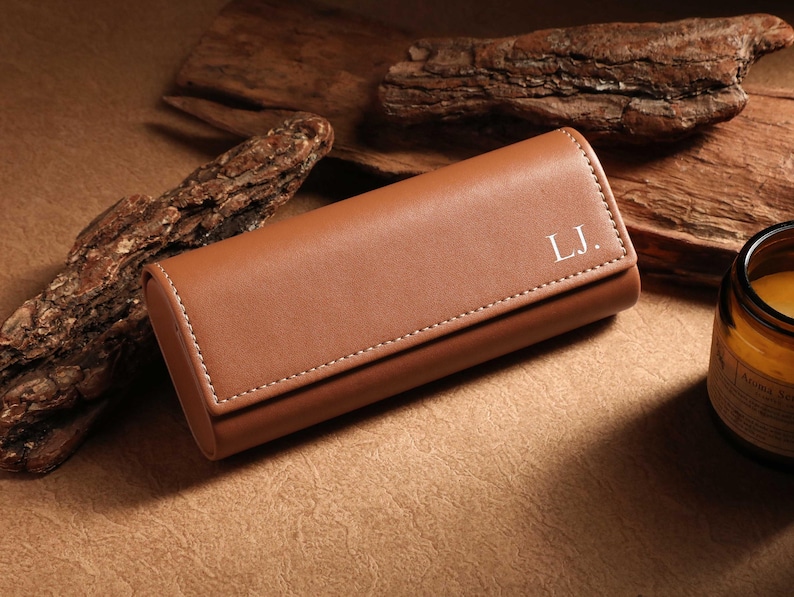 May include: A brown leather glasses case with white stitching and the initials "L.J." in white. The case is rectangular with rounded edges and a flap closure. The background includes wood pieces and a candle.