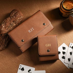 May include: Two brown leather cases, one rectangular and one square, with silver and gold initials. Playing cards with black spade symbols are scattered around the cases. A candle and a jar are also in the image.