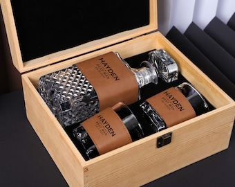 May include: A wooden box with a brown leather lid, containing two glass decanters with brown leather wraps. The leather wraps are engraved with the text "Hayden Best Man 2023".