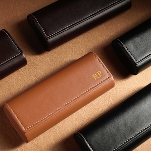 Peut inclure: Plusieurs étuis à lunettes en cuir de différentes couleurs : marron, noir et beige. Chaque étui est rectangulaire avec des bords arrondis et des coutures blanches. Un étui beige porte les initiales dorées "RP".