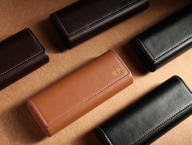 May include: An assortment of leather eyeglass cases in various colors: brown, black, and tan. Each case is rectangular with rounded edges and white stitching. One tan case features the gold initials "RP".