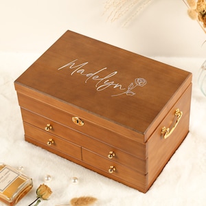 May include: A wooden jewellery box with a personalised inscription "Madelyn" and a delicate rose design. The box has four drawers with gold-coloured handles and a lock.