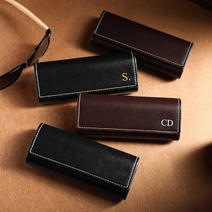 May include: An assortment of leather eyeglass cases in black and brown, with white stitching. Some cases have initials in gold or white. Sunglasses with light-colored arms are also present, along with a lit candle in a jar.