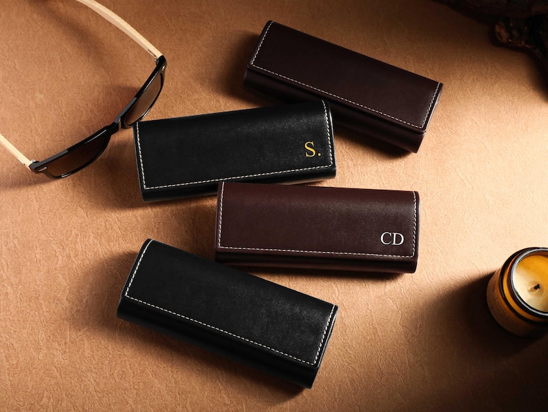 May include: An assortment of leather eyeglass cases in black and brown, with white stitching. Some cases have initials in gold or white. Sunglasses with light-colored arms are also present, along with a lit candle in a jar.