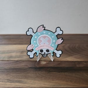 May include: Embroidered patch featuring a skull and crossbones design with a blue and pink color scheme. The patch has a circular center with an X, surrounded by a light blue border and pink accents. The skull has a black outline.