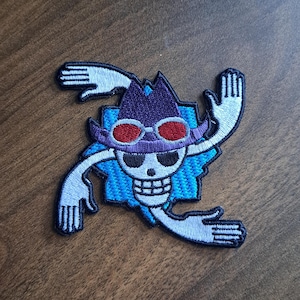 May include: Embroidered patch depicting a skull with red goggles, a purple hat, and outstretched arms. The design is set against a blue background with a white outline. Suitable for application on clothing or accessories.