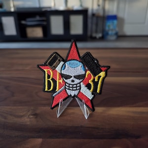 May include: An embroidered patch featuring a red star background with a central skull icon wearing blue hair and black sunglasses. The design includes two crossed black tools and the yellow text BF 37, all outlined in black thread on a fabric base.