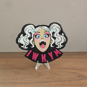 May include: An embroidered patch featuring a cartoon character with white pigtails, blue eyes, and fangs, set against a black background. The character has pink eyeshadow and an open mouth. The text IWKYM is stitched in pink letters at the bottom.