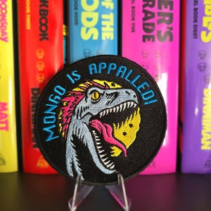 May include: A circular embroidered patch featuring a light blue dinosaur head with a pink tongue, yellow accents, and the text "MONGO IS APPALED!" in blue lettering against a black background. The patch is displayed on a clear plastic stand.