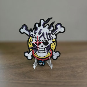 Puede incluir: Un parche bordado que presenta una calavera estilizada con cabello blanco, una cruz roja sobre un ojo y un fondo circular amarillo con marcas negras. El diseño incluye huesos cruzados, un collar de cuentas rojas y detalles morados.