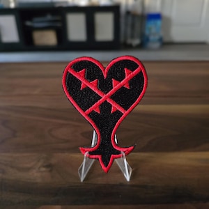 May include: A heart-shaped embroidered patch featuring a black base with a vibrant red border. The design includes two intersecting red lines that cross through the center of the heart, creating a sharp, angular pattern against the black fabric.