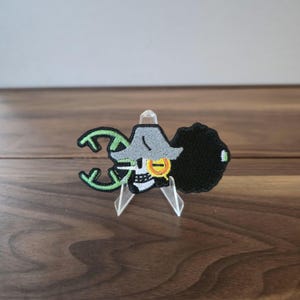 Puede incluir: Parche bordado con una calavera que lleva un sombrero de pirata gris y un afro negro. El parche tiene detalles verdes y dorados. La calavera tiene un emblema dorado. El parche se muestra sobre un soporte transparente.