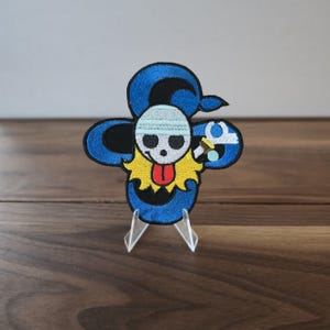 May include: Embroidered patch featuring a skull with a blue hat and a yellow collar. The patch has a blue outline and a black background. The skull has a red tongue and is holding a small object. The patch is displayed on a clear stand.